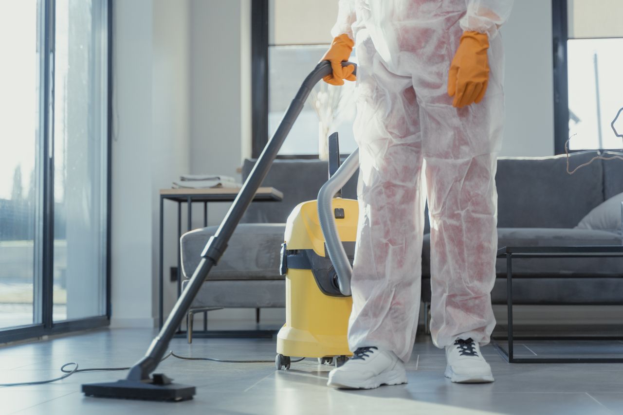 services-01 A cleaner in protective coveralls and gloves vacuuming a modern living room.