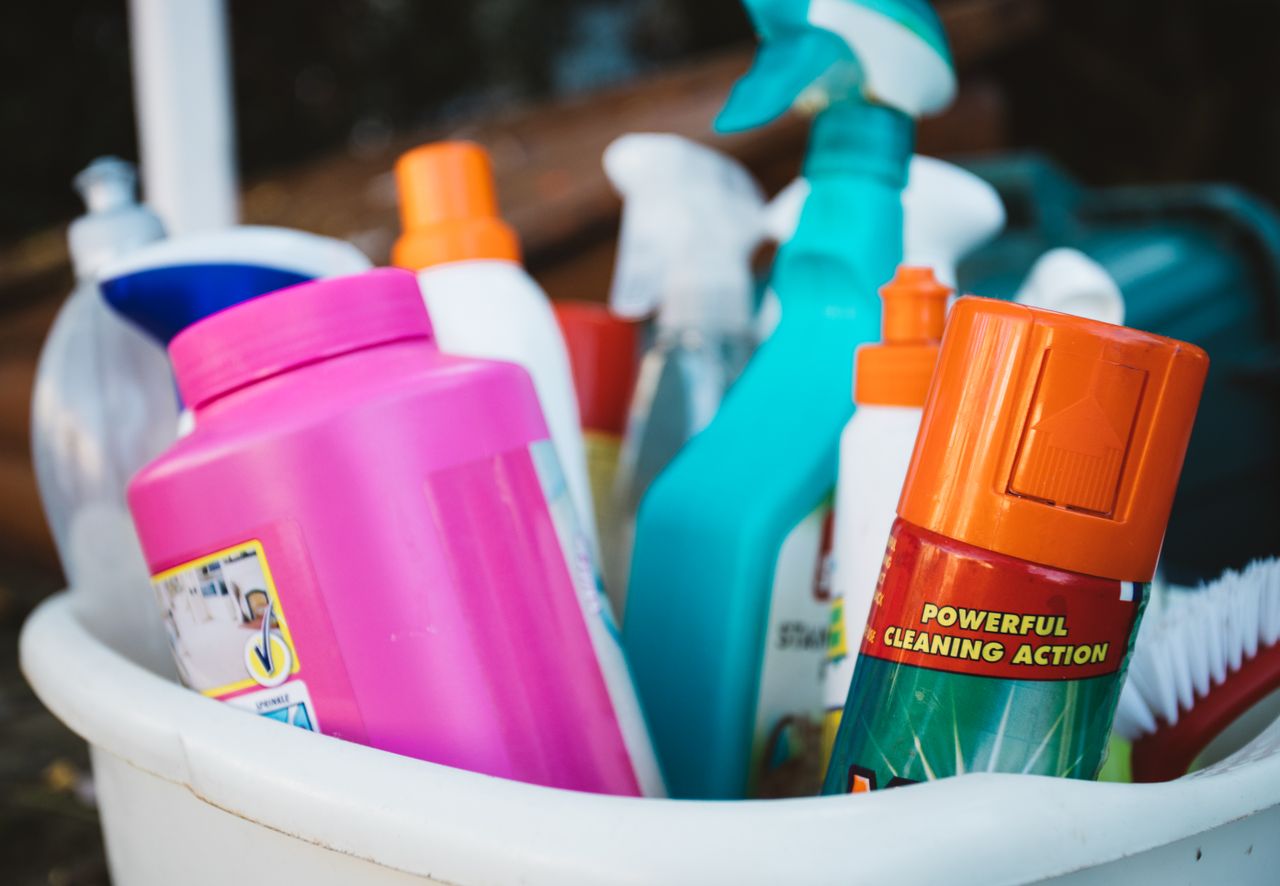 hero-img-02 Close-up of assorted cleaning supplies in a bucket outdoors, showcasing various detergents and sprays.